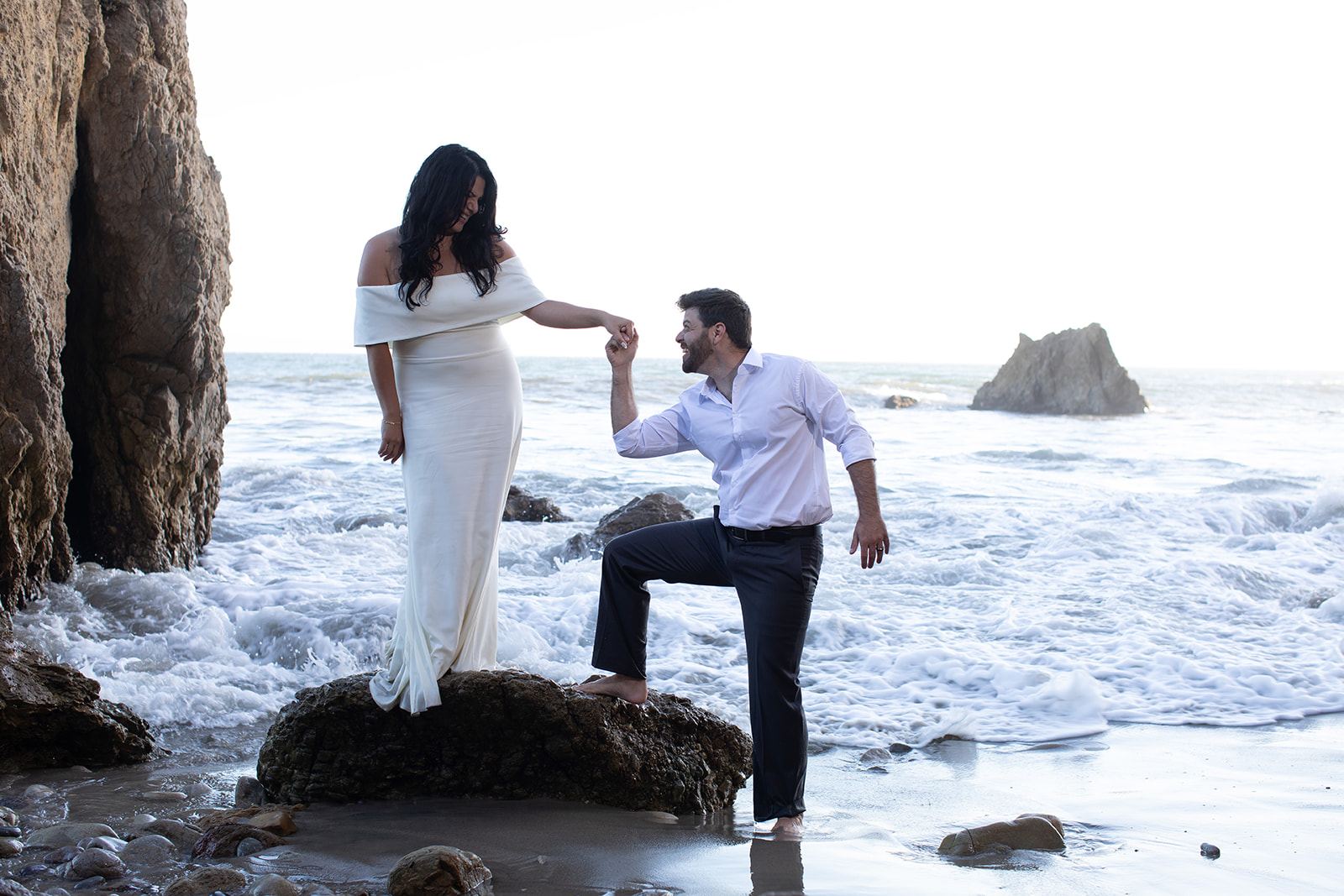 Sari kneeling on a rock to kiss Cam's hand at the water's edge