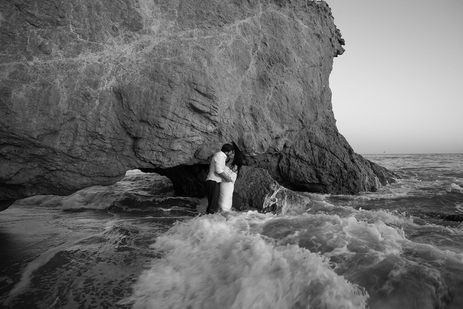 Cam and Sari kissing beneath a sea cliff as waves crash around them